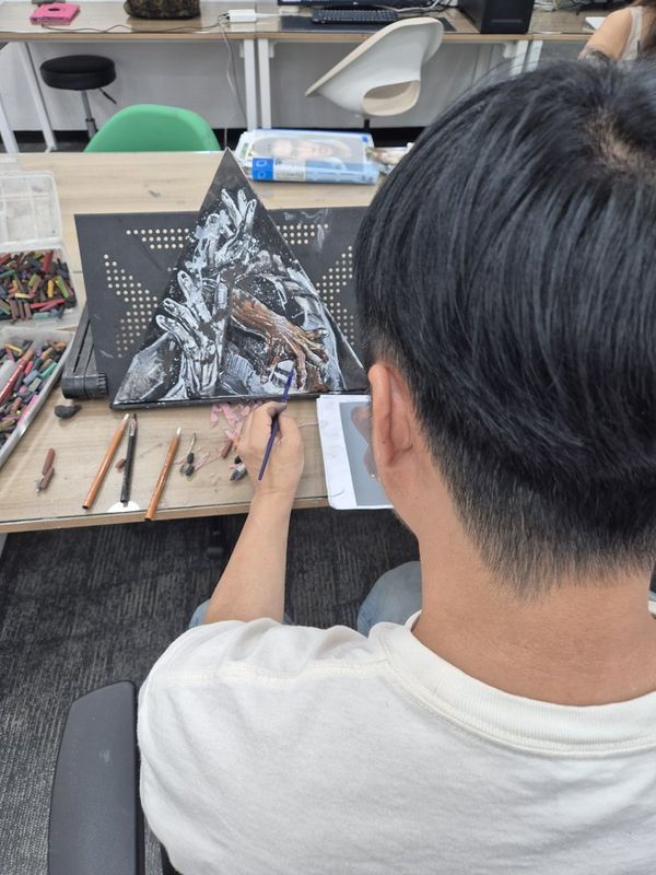 Close-up photograph of a student's hand working on a detailed dark mixed-media artwork in a Royal Blue studio, with brushes and colorful references visible on the table