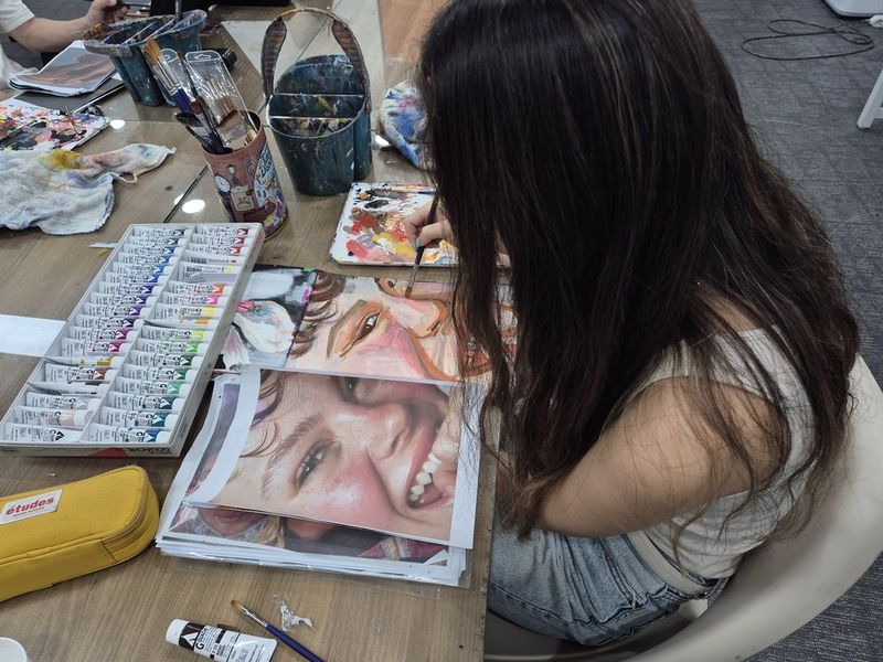 Photograph of a student working at a Royal Blue studio desk surrounded by colorful artworks and references, using colored pencils or markers on a detailed illustration