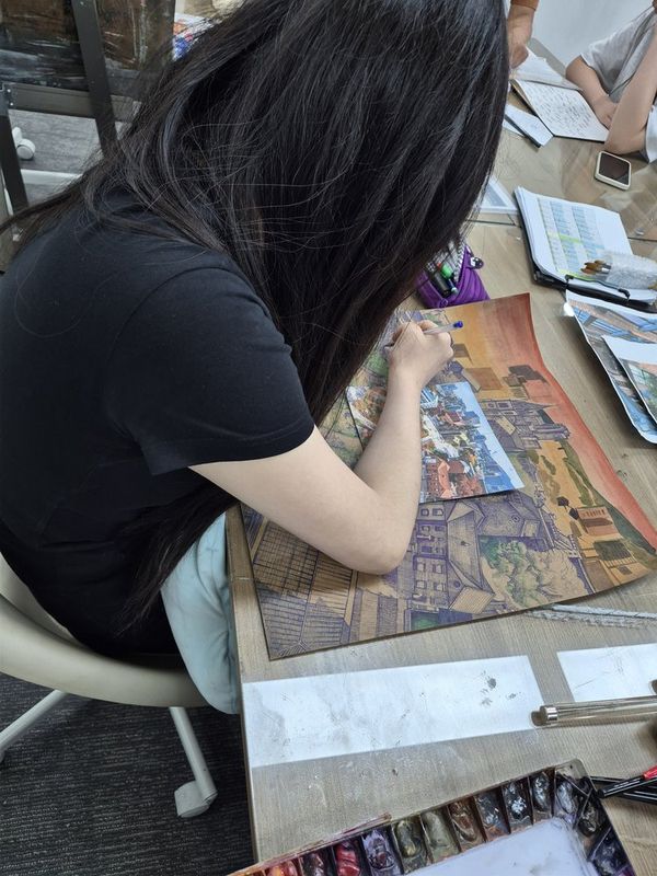 Close-up photograph of a student's hands working on a detailed portrait or figurative drawing at a Royal Blue studio table, with pencils and reference visible on the surface