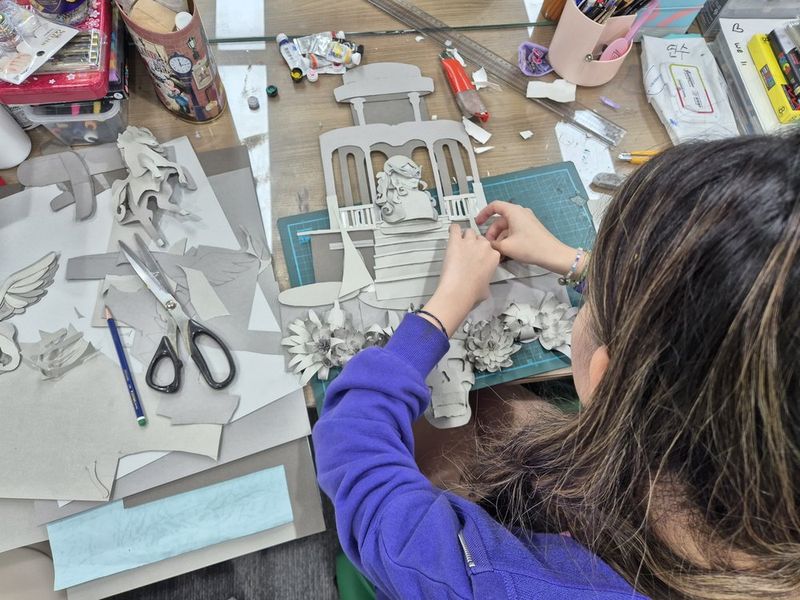 Close-up overhead photograph of a student's hands working on a sketch or outline drawing on white paper at a Royal Blue studio, with colored pencils visible nearby