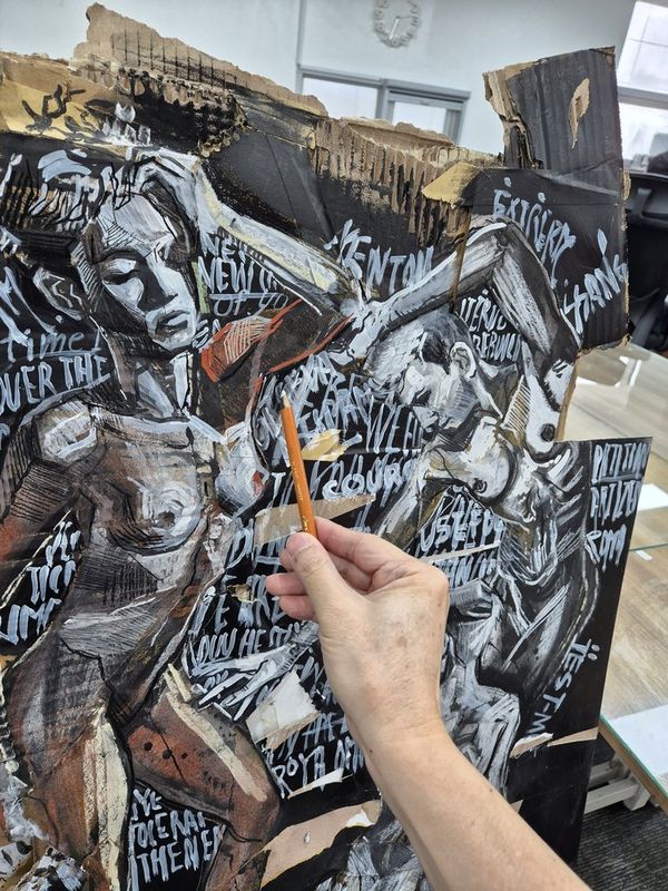Photograph showing an instructor's hands adding detail to a large dramatic black-and-white mixed-media artwork in a Royal Blue studio, surrounded by reference imagery and art supplies