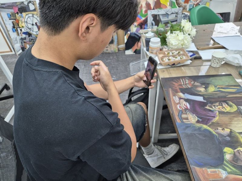 Overhead close-up photograph of a student working on a detailed drawing or illustration at a Royal Blue studio desk, showing the artwork in progress with dark tonal shading
