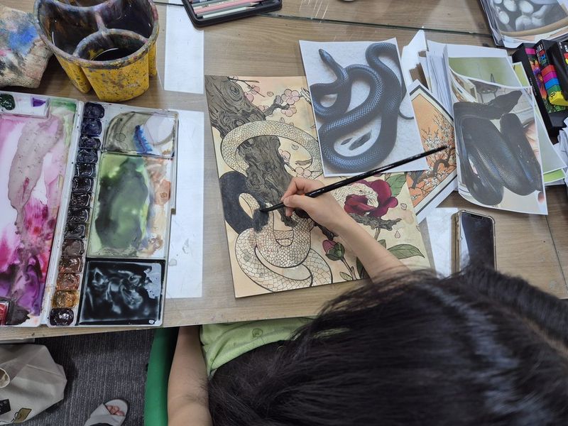 Overhead photograph of a Royal Blue studio work surface showing a student working on dark mixed-media artworks with brushes and reference images spread on the desk