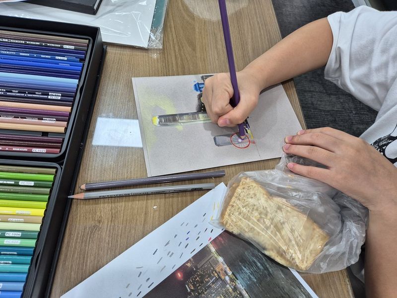 Close-up photograph of a student's hands working with colored pencils or pastels on a detailed artwork at a Royal Blue studio, with a pencil case and art supplies visible nearby