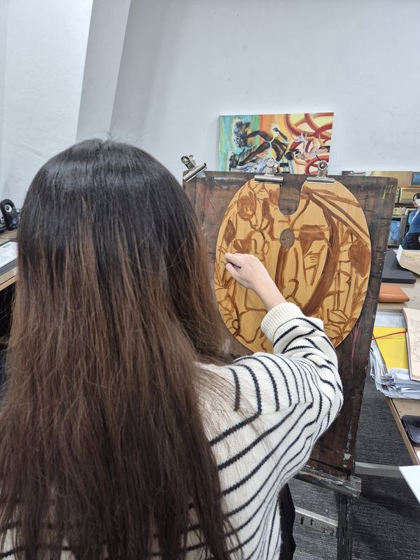 Photograph of a student working on a large detailed painting at a Royal Blue studio, viewed from behind, showing the student engaged with a warm-toned figurative artwork on canvas