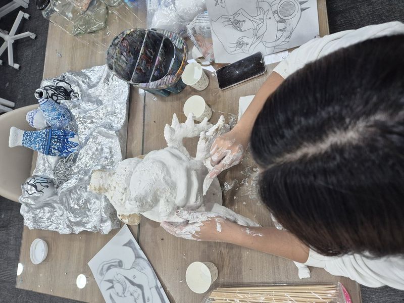 Photograph of a student at a Royal Blue studio working on a three-dimensional sculptural or ceramic project with white material, with studio shelves and art supplies visible in the background