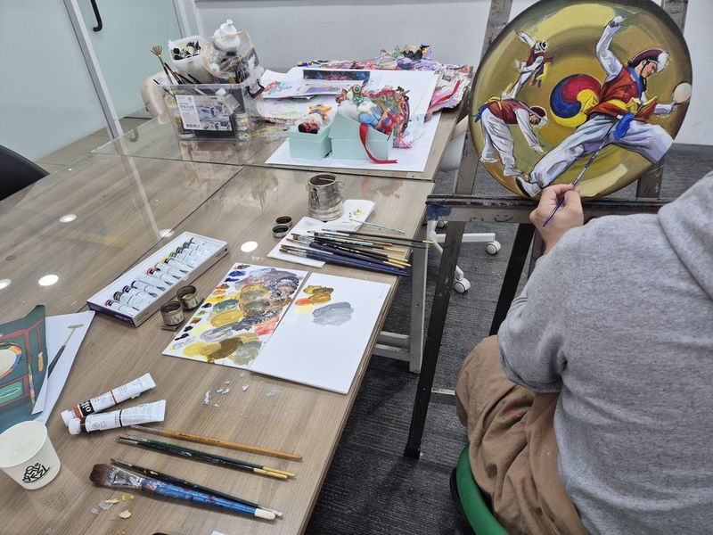 Photograph of a Royal Blue studio class in session showing a student working at a desk on a detailed artwork, with studio equipment and other students visible in the background