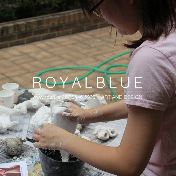 Royal Blue branded close-up photograph of a student's hands sculpting or modeling a three-dimensional form using white clay or sculptural material at a studio table