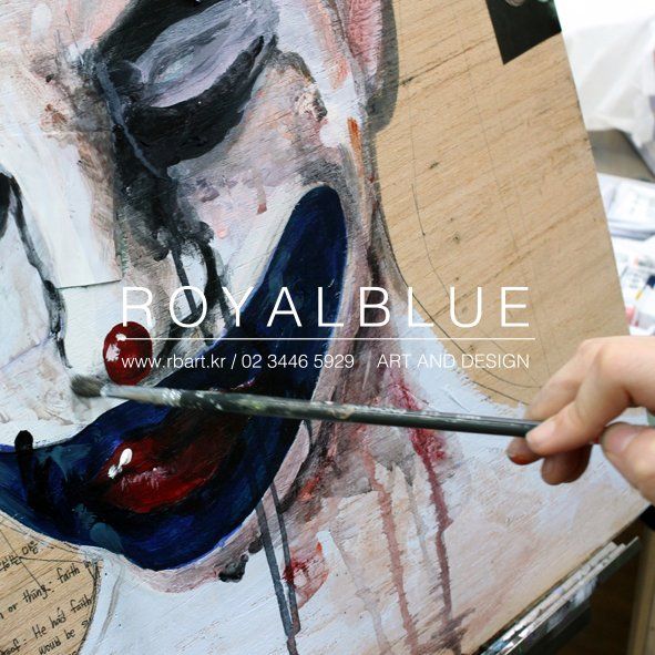 Royal Blue branded close-up overhead photograph showing hands painting a dark dramatic mixed-media artwork on a canvas, with bold gestural marks and reference imagery around the workspace