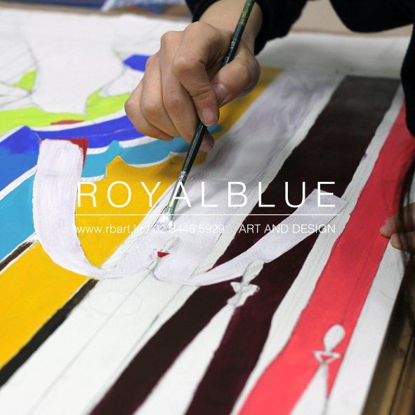 Royal Blue branded close-up photograph of a student's hands working with large paint brushes on a colorful geometric or abstract artwork, showing bold diagonal strokes in multiple colors