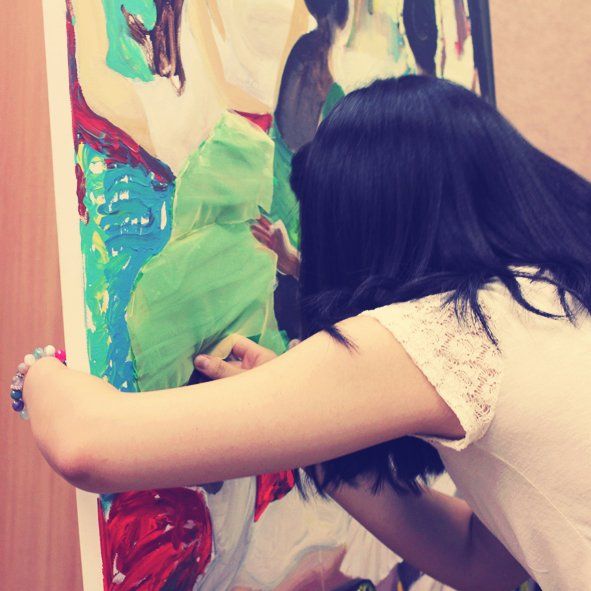 Royal Blue branded photograph of a student working on a large canvas mounted on an easel, applying paint with broad brushstrokes to a colorful artwork in a Royal Blue studio