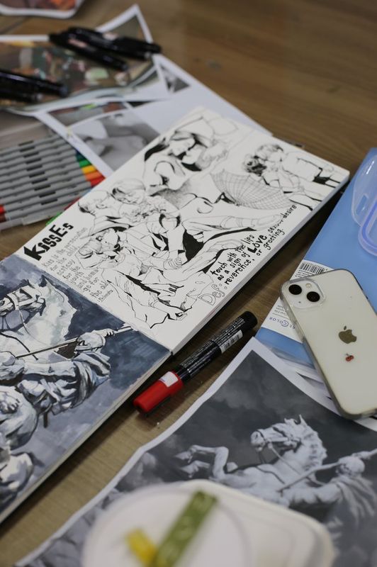 Close-up overhead photograph of a student working on a black-and-white manga or comic illustration with fine pen lines on a drawing surface at a Royal Blue studio