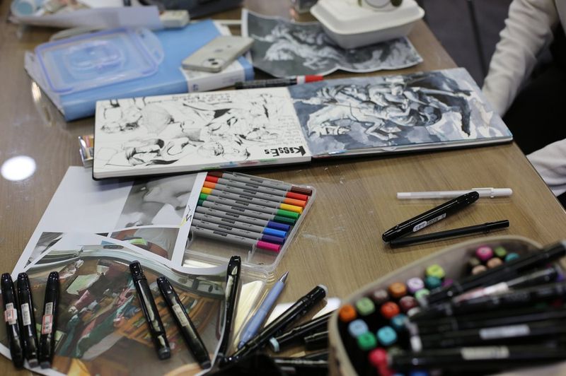 Overhead photograph of a Royal Blue studio desk showing black-and-white comic or manga illustration work in progress, with ink pens and markers spread around the drawing surface
