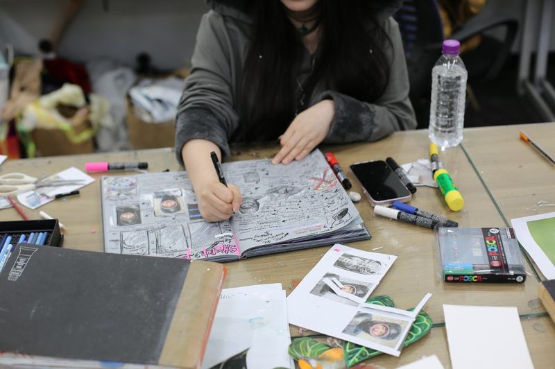 Photograph of a student at a Royal Blue studio table working on a detailed drawing or illustration, viewed from behind, with a water bottle and art supplies visible nearby