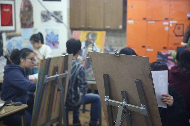Interior photograph of a Royal Blue studio class in session with multiple students working on easel paintings at a large open studio space with artwork visible on walls