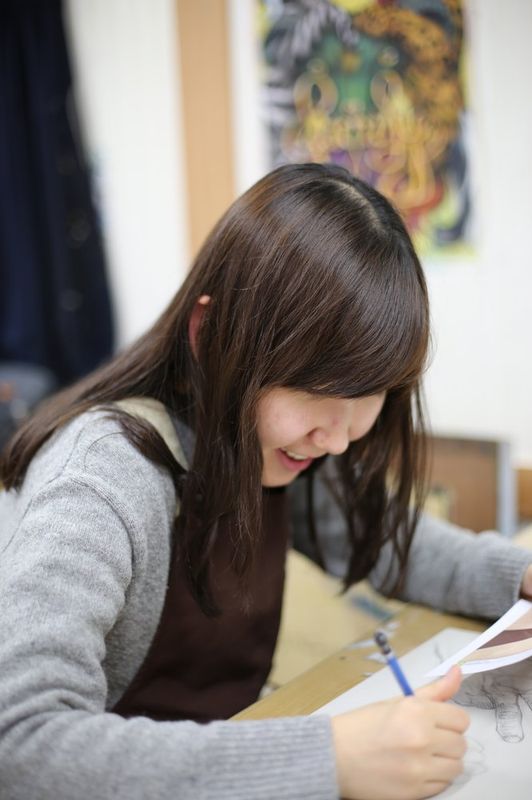 Photograph of a female student at a Royal Blue studio smiling while working on a drawing or illustration at a desk, with artworks visible in the background