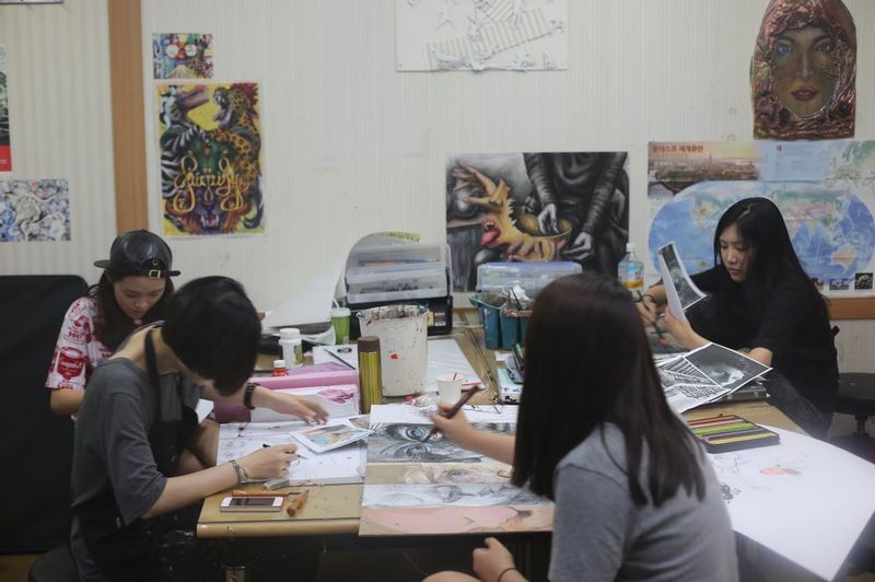 Wide photograph of a Royal Blue studio class showing two female students working at a table surrounded by colorful artworks, with other students and art supplies visible in the background