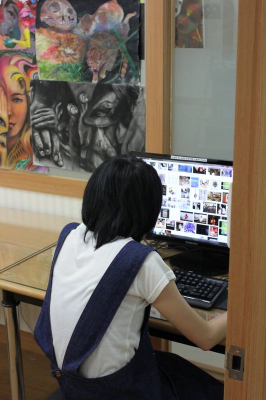 Photograph of a student at a Royal Blue studio looking at a laptop screen with reference images, working on a digital design or illustration project at a desk surrounded by art materials