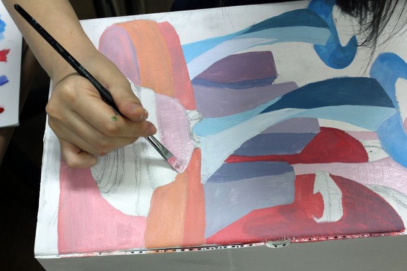 Close-up photograph of a student's hand using a wide brush to apply bold strokes of paint to a large colorful abstract or figurative artwork in a Royal Blue studio