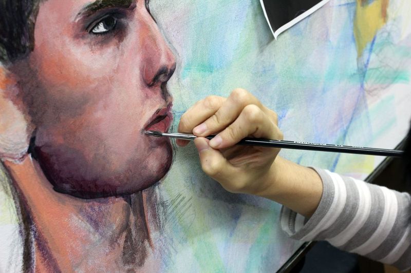 Close-up photograph of an instructor or student's hands painting a hyper-realistic close-up portrait face with a fine brush, showing precise application of paint on a large canvas