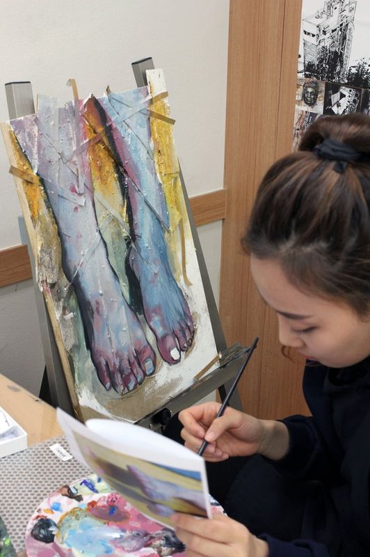 Photograph of a student at a Royal Blue studio standing before a large canvas mounted on an easel, working on a detailed figurative painting with a brush