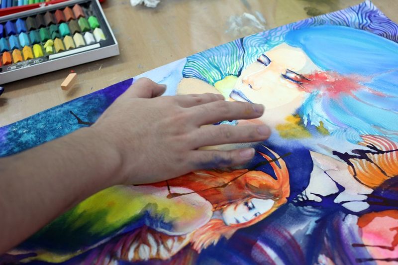Close-up photograph of a student's hand painting detailed work on a colorful figurative artwork at a Royal Blue studio, showing careful brushwork on an anime or illustration-style face