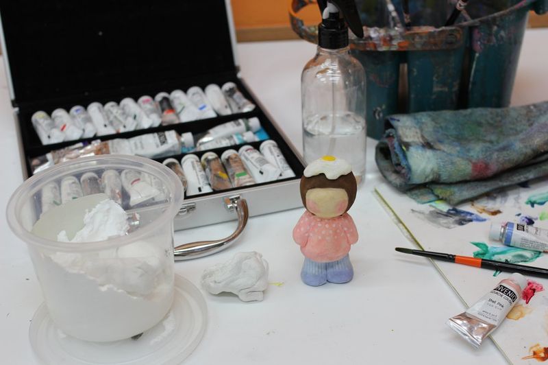 Close-up overhead photograph of a Royal Blue studio work surface showing art supplies including paint containers, small sculptural objects, and various mixed media tools for a class project