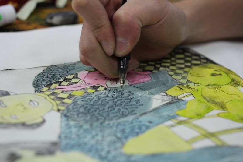 Close-up overhead photograph of a student's hands carefully working with a fine pen on a detailed mixed-media artwork combining photographic cutouts and drawn elements on yellow and green tones