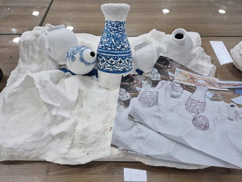 Overhead photograph of a Royal Blue studio desk or surface showing white fabric or textile pieces alongside a small decorative ceramic or glass object and art materials