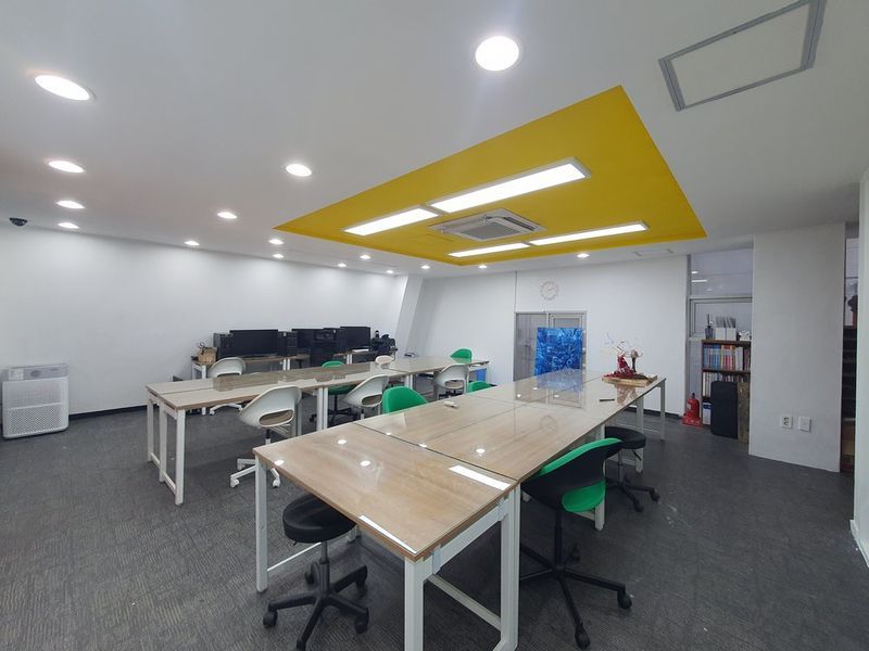 Interior photograph of a modern art studio classroom with white desks, bright overhead lighting, yellow ceiling details, and colorful artwork displayed on the walls