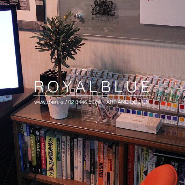 A Royal Blue branded interior photograph showing a cozy studio reading nook or bookshelf corner with a small plant, books, and warm lighting with studio watermark overlay