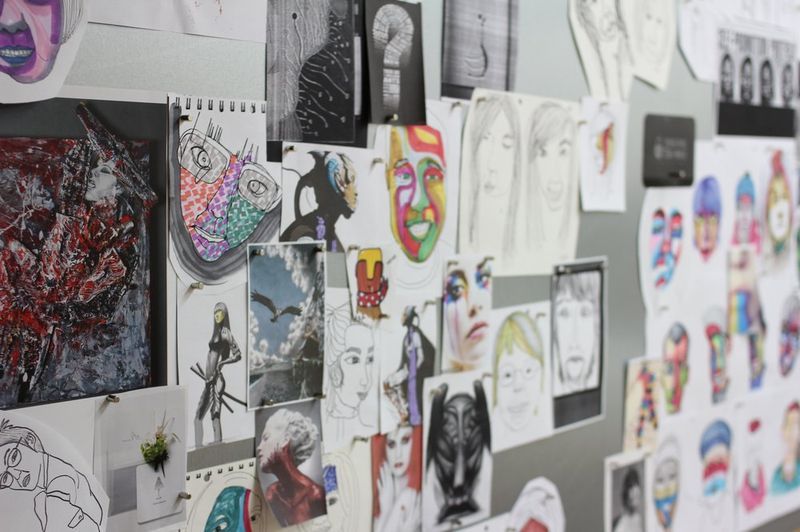 Interior photograph of a Royal Blue studio wall or bulletin board covered with fashion illustration sketches, character designs, and colorful artwork prints in an organized display