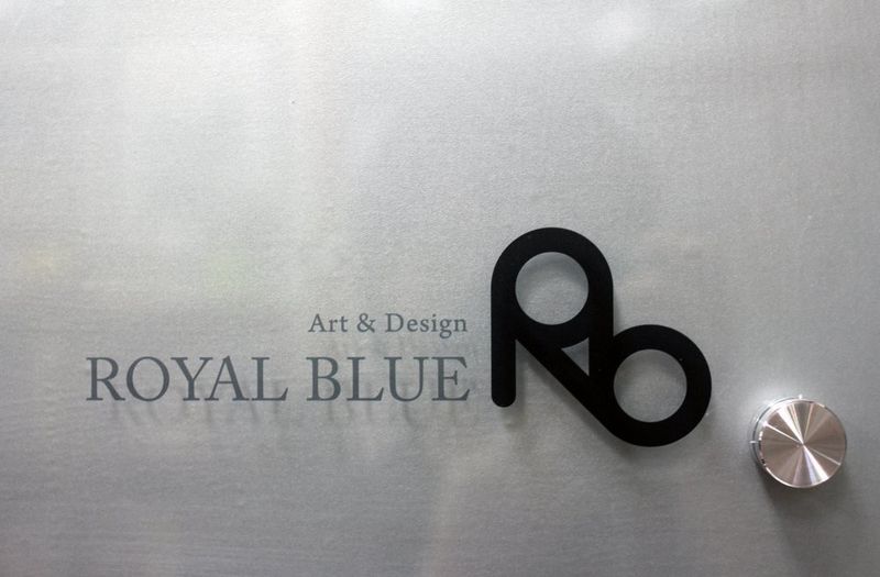 Close-up photograph of the Royal Blue Art and Design studio entrance sign featuring the Rb logo mark and full studio name in black lettering on a brushed metal or frosted surface