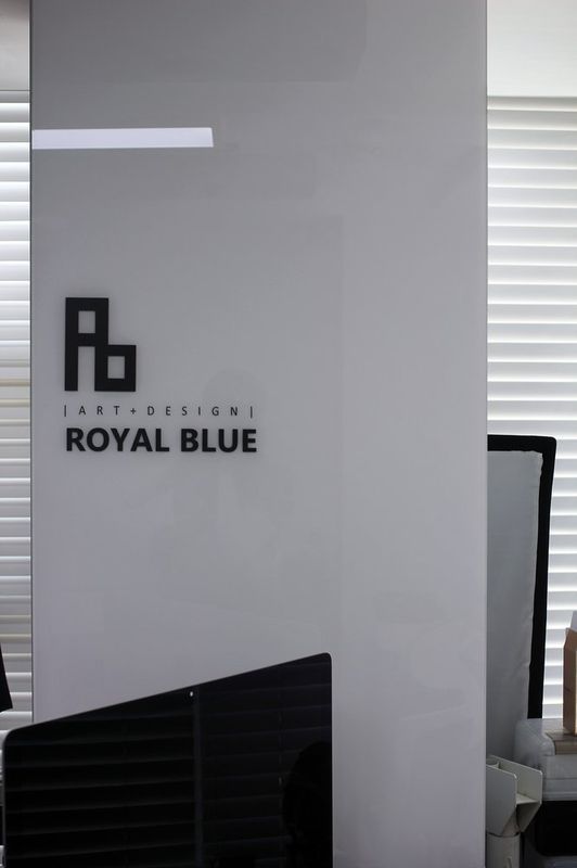 Interior photograph of a Royal Blue studio entrance area showing the Rb logo displayed on a wall panel with a black sofa visible in the corridor leading to studio rooms