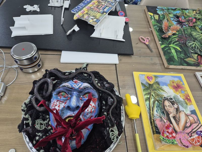 Photograph of a Royal Blue studio work surface showing student artworks spread out including a dark figurative painting with blue tones and various drawings under review