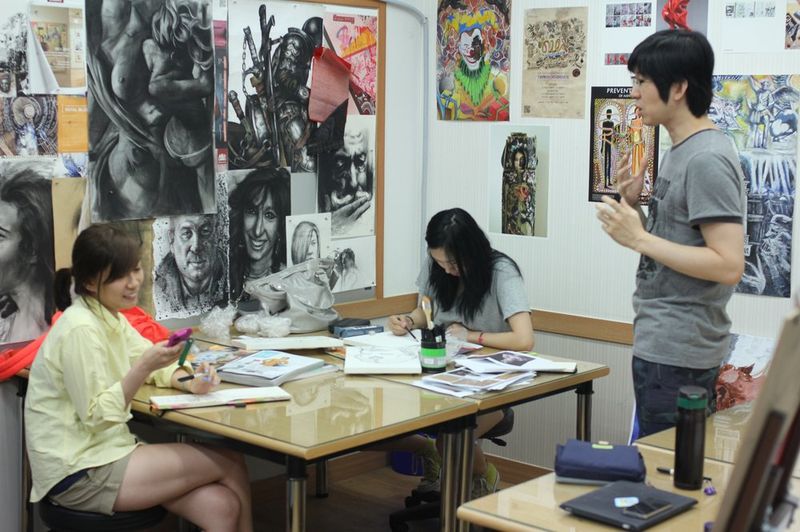 Photograph of a Royal Blue studio class session showing students working at tables with artwork and reference materials visible, in a bright studio environment with black-and-white art on the walls