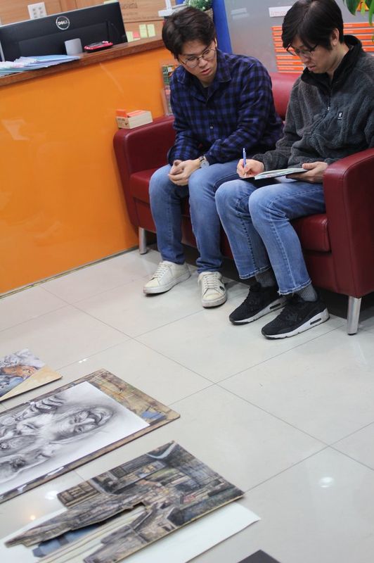 Photograph of a Royal Blue studio group session showing three people seated on chairs in a circle in a review or consultation meeting, with large artworks displayed on the floor around them